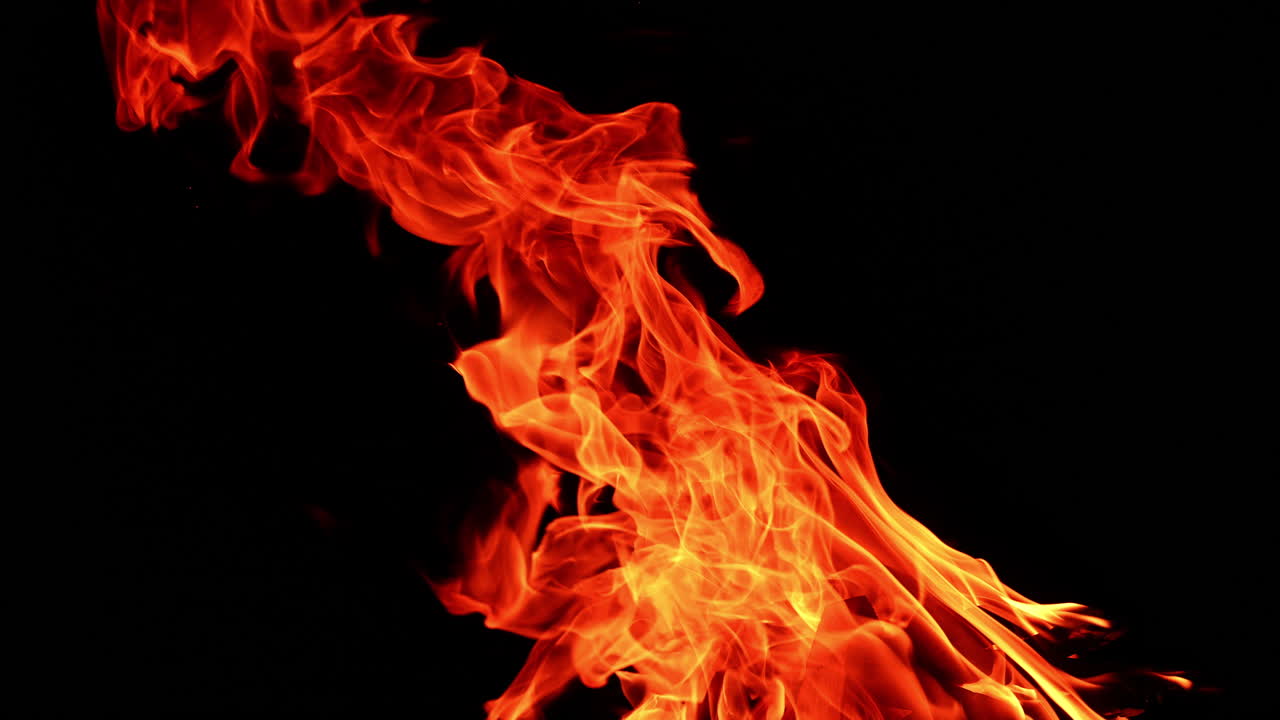 Pieces of wood burning in the big bonfire. Long flame stretches into the air leaning to the left. Darkness at backdrop.