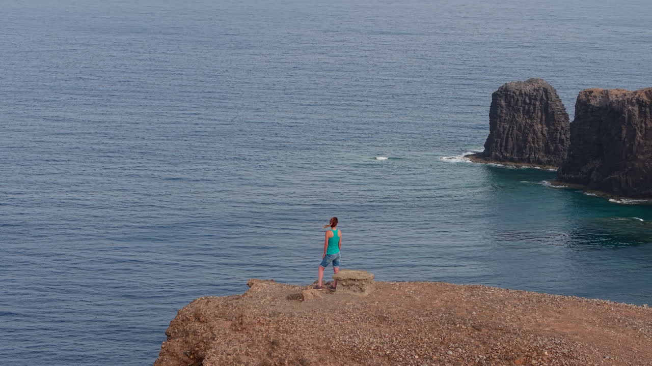 Female traveler standing on rocky cliff edge, gazing at iconic Roque Partido volcanic landmark with expansive ocean panorama stretching across Gran Canaria landscape