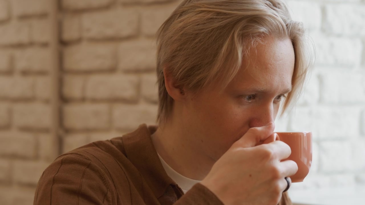Primo piano di un giovane uomo che beve da una tazza di ceramica rosa davanti a un muro di mattoni bianchi. Il soggetto tiene la tazza con entrambe le mani creando un momento intimo contro una luce naturale soffusa. Dettagli dello sfondo.