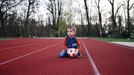 lovely baby boy sits on the stadium track holding a ball. calm caucasian toddler looks around and stands up.