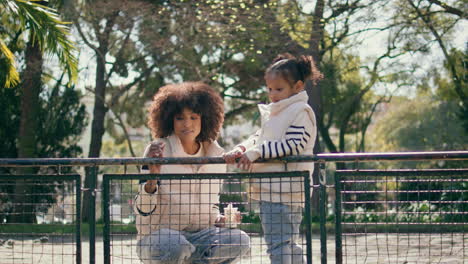 mother daughter standing park fence sunny day. mom throwing breadcrumbs