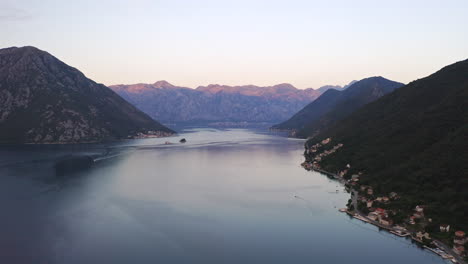 The-bay-of-Kotor-in-Montenegro-with-a-coastal-town-on-its-shore,-mountain-ranges-surrounding-the-bay-and-dominating-the-horizon,-cloudless-sky-above,-red-morning-glow-shining-on-its-peaks,-aerial