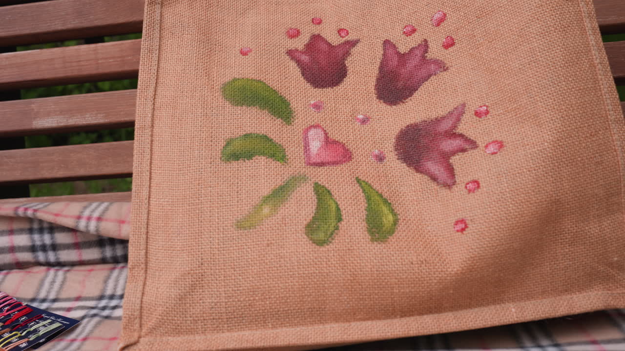 Primer plano de un bolso de mano pintado con un diseño floral y de corazones sobre una manta de cuadros junto a hilos de bordar de colores, tijeras y un aro, mostrando arte creativo hecho a mano.