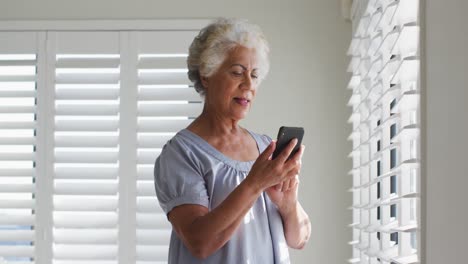 Mujer-Mayor-Afroamericana-Usando-Un-Teléfono-Inteligente-Y-Mirando-Por-La-Ventana-En-Casa