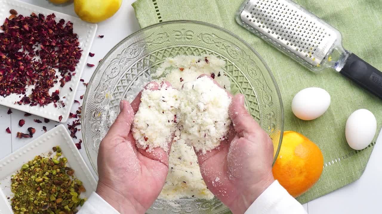 Baking a dessert called Persian Love Cake made with rose petals and pistachios. Cake with white glaze frosting. Using hands to mix in the ingredients.. Beautiful cake for tea time or celebration