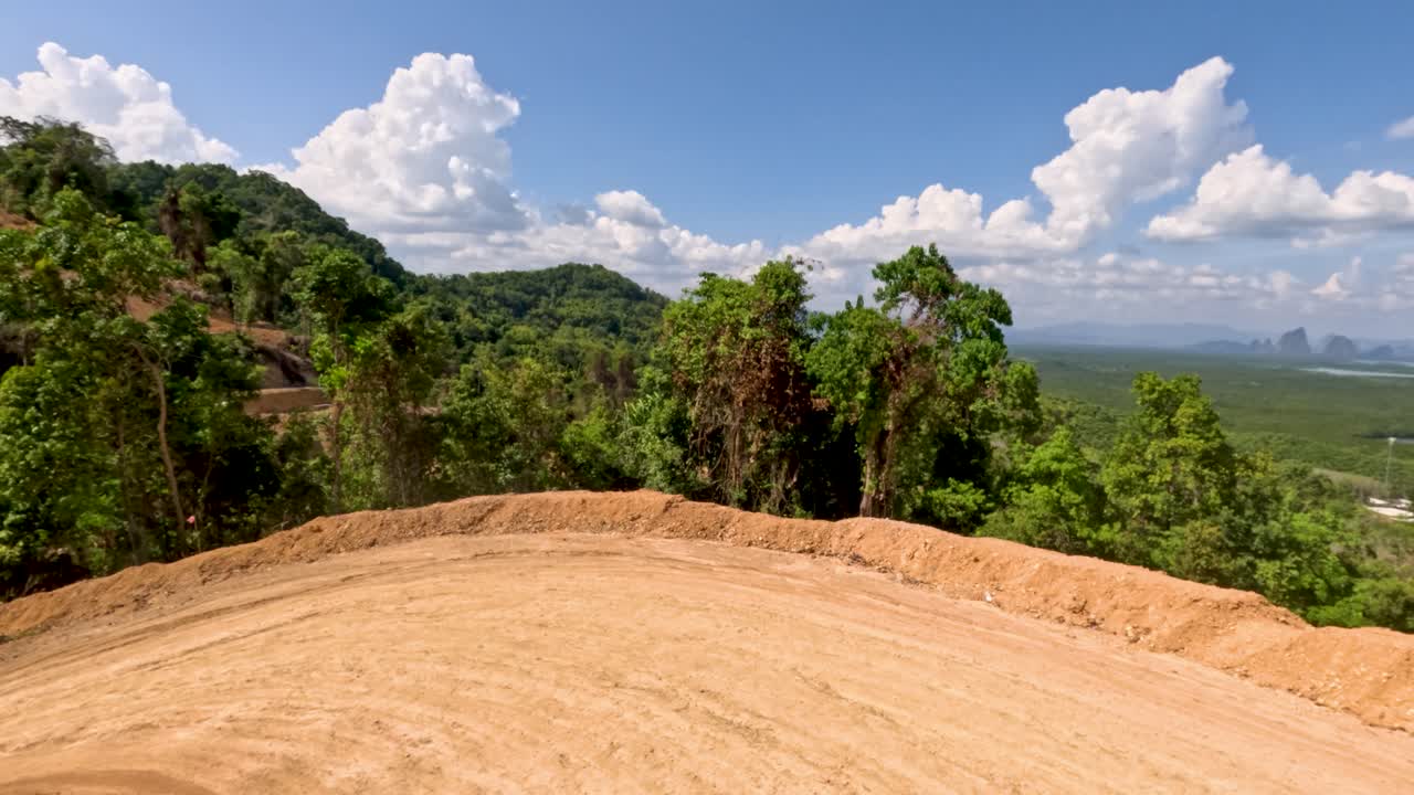 Camera moves along sunlit dirt road through tropical hillside landscape, lush greenery, and distant mountains