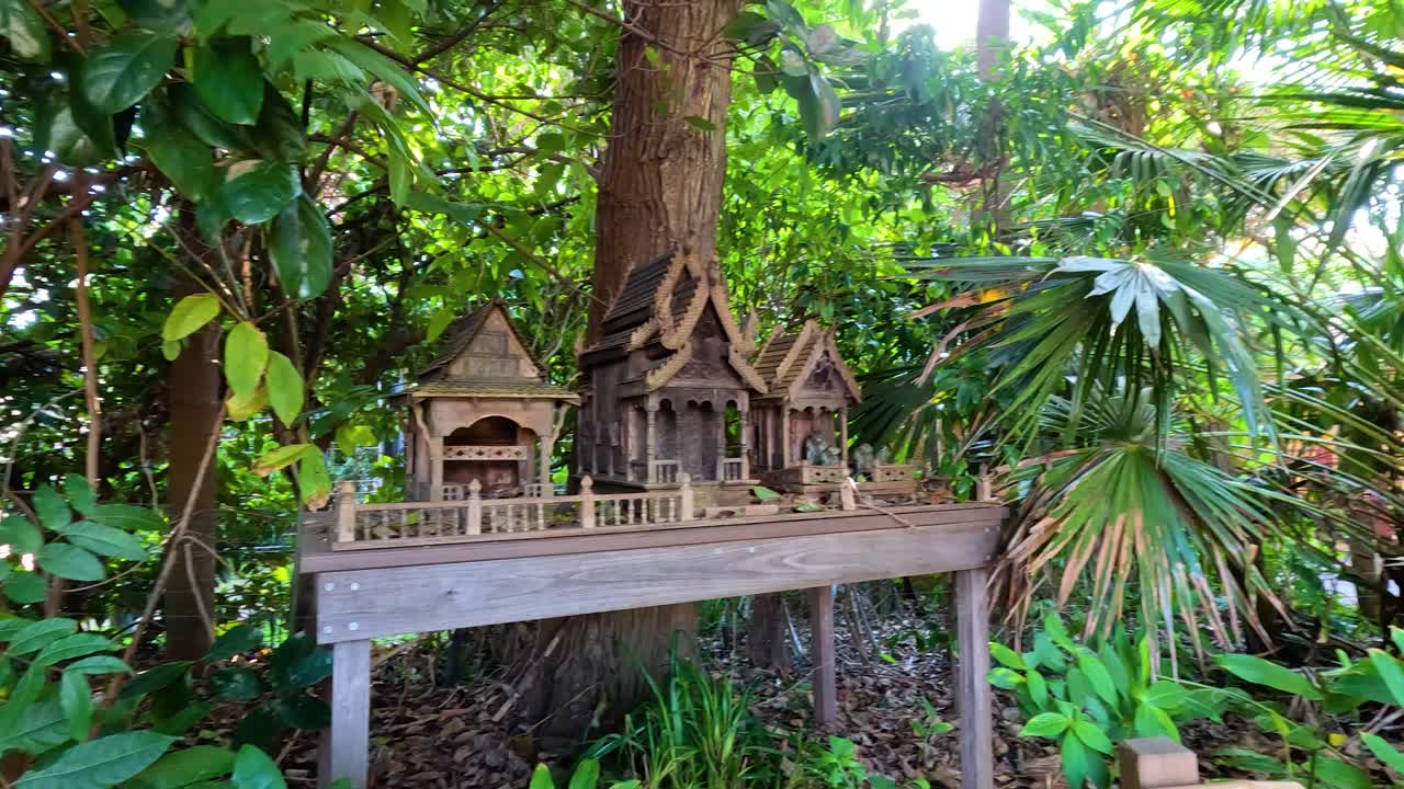 잔잔한 초록색으로 둘러싸인 전통적인 태국 정신의 집을 특징으로하는 조용한 정원 장면, 부드러운 카메라 움직임과 자연 조명으로 포착