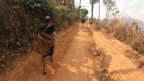 Un-Anciano-Asiático-Con-Una-Cesta-Vacía-Caminando-Por-Una-Pequeña-Carretera-En-Las-Montañas-De-Tailandia,-Con-Otras-Personas-En-El-Fondo