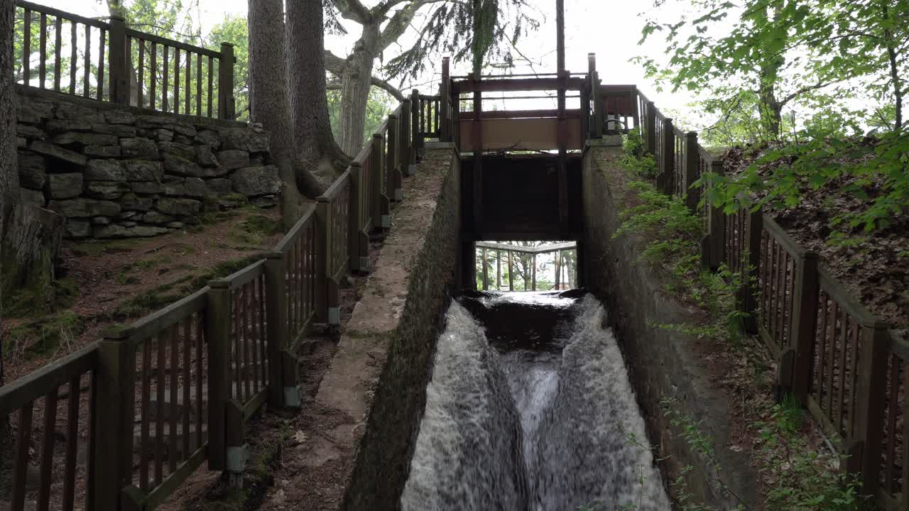 A waterfall flowing with a strong current from a bridge surrounded by concrete on both sides. Around it, there are trees with some greenery.