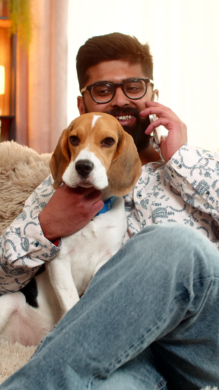Indian man talks on smartphone while sitting on sofa with beagle dog next to him at home indoors