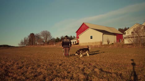 Man-Pushing-Wheelbarrow-With-Alaskan-Malamute-Dog-Breed-In-Countryside-Village