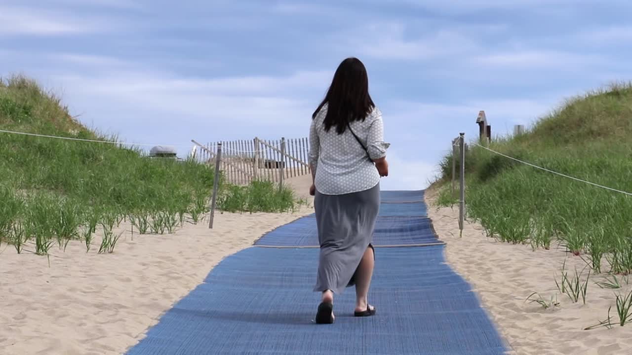 A woman is walking away from the camera at a beach with a blue mat placed on the sand.