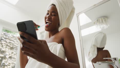 Video-Einer-Glücklichen-Afroamerikanischen-Frau-In-Robe,-Die-Zähne-Putzt-Und-Ihr-Smartphone-Benutzt