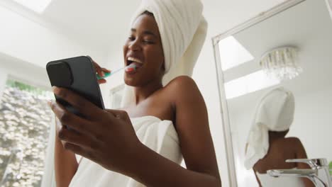 video de una feliz mujer afroamericana en túnica cepillándose los dientes y usando un teléfono inteligente
