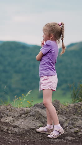 cute little girl in purple clothes thinks about travelling touching cheek by finger on hill against old mountains at eco resort on gloomy day slow motion