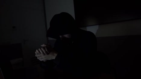 Close-up-of-ominous-criminal-caucasian-male-wearing-black-hoodie-while-sitting-in-at-desk-in-darkness