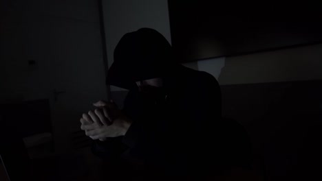 close up of ominous criminal caucasian male wearing black hoodie while sitting in at desk in darkness