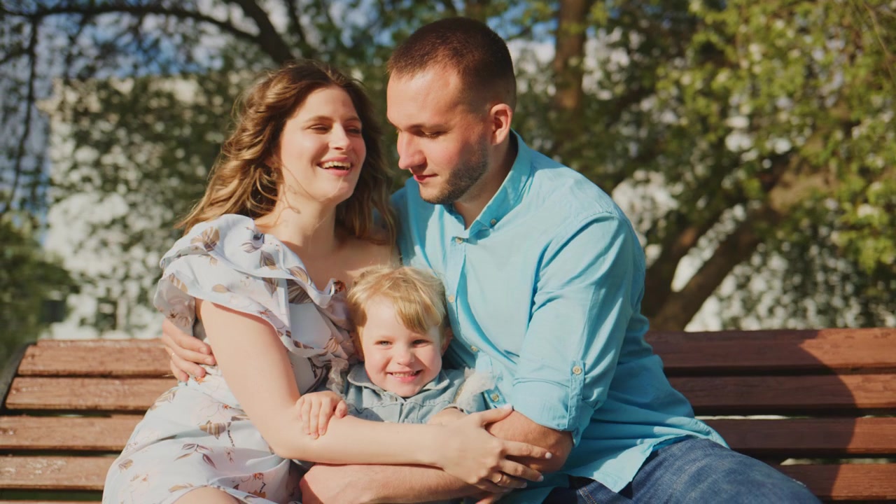 Famiglia felice di tre persone su una panchina del parco. La madre abbraccia il bambino mentre il padre guarda con amore. Il bambino sorride gioiosamente tra i genitori. La luce calda e naturale esalta il momento di pace. Ideale per famiglia, genitorialità.