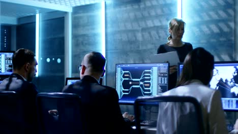 in the government system control center. female supervisor with laptop holds meeting for her team of engineers. room is equipped with multiple displays showing various information.