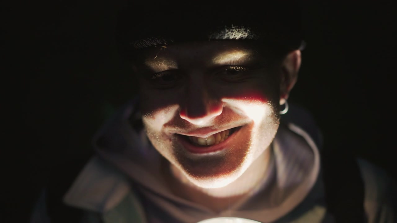 closeup flashlightlit caucasian face smiling, camp storyteller shifts roles teen camper, trail guide, urban explorer, latenight poet, prankster warm glow from handheld lantern illuminates hoodie