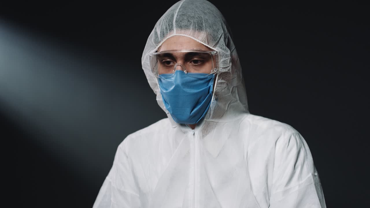 médico varón caucásico con equipo de protección personal y máscara médica, poniéndose anteojos y mirando al lado sobre fondo de pared negra