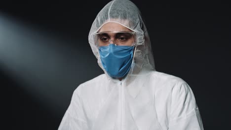médico varón caucásico con equipo de protección personal y máscara médica, poniéndose anteojos y mirando al lado sobre fondo de pared negra
