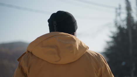 round view wildlife photographer wearing yellow winter coat beanie gloves standing on snowy street spot looking around sipping drink holding camera against blur city buildings under winter daylight
