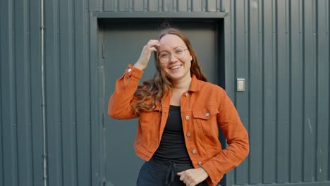 Una-Joven-Sorprendida-Con-Una-Vibrante-Chaqueta-Naranja-Doblando-Los-Brazos-Muestra-Una-Encantadora-Felicidad-De-Emociones-Contra-Una-Pared-Gris