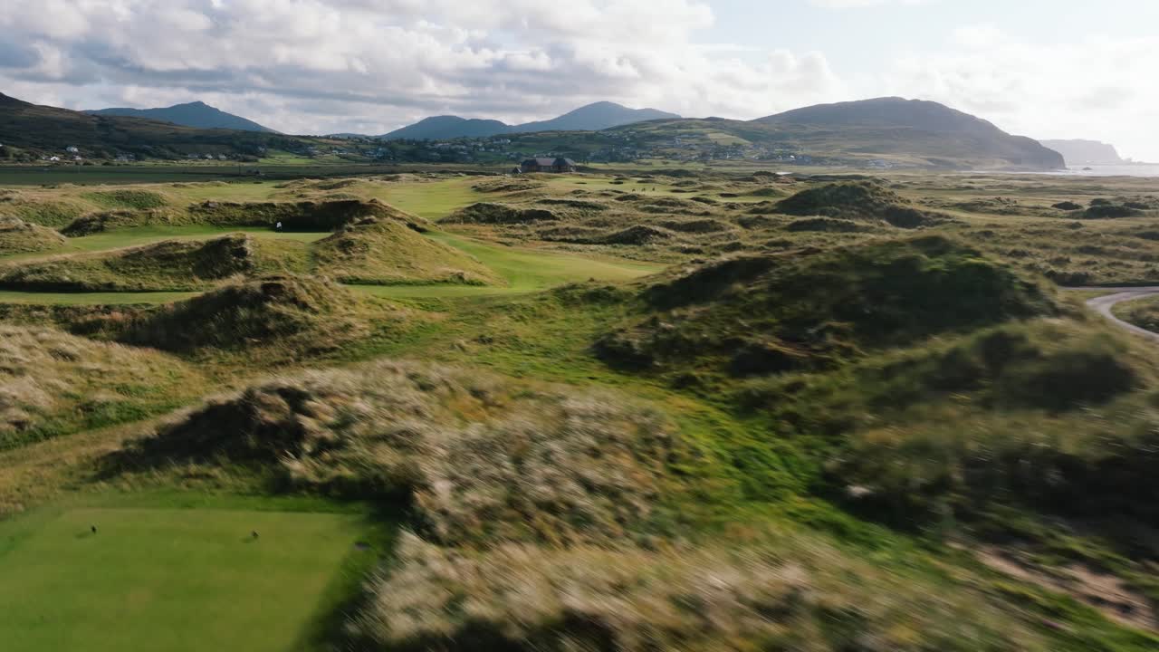Sorvolo con drone di colline verdi ondulate, fairway e bunker su un classico campo da golf links in Irlanda, che termina alla clubhouse con vista sull'oceano.