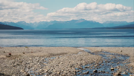 Small-stream-flowing-across-a-rocky-beach-into-a-tranquil-lake-surrounded-by-distant-mountains