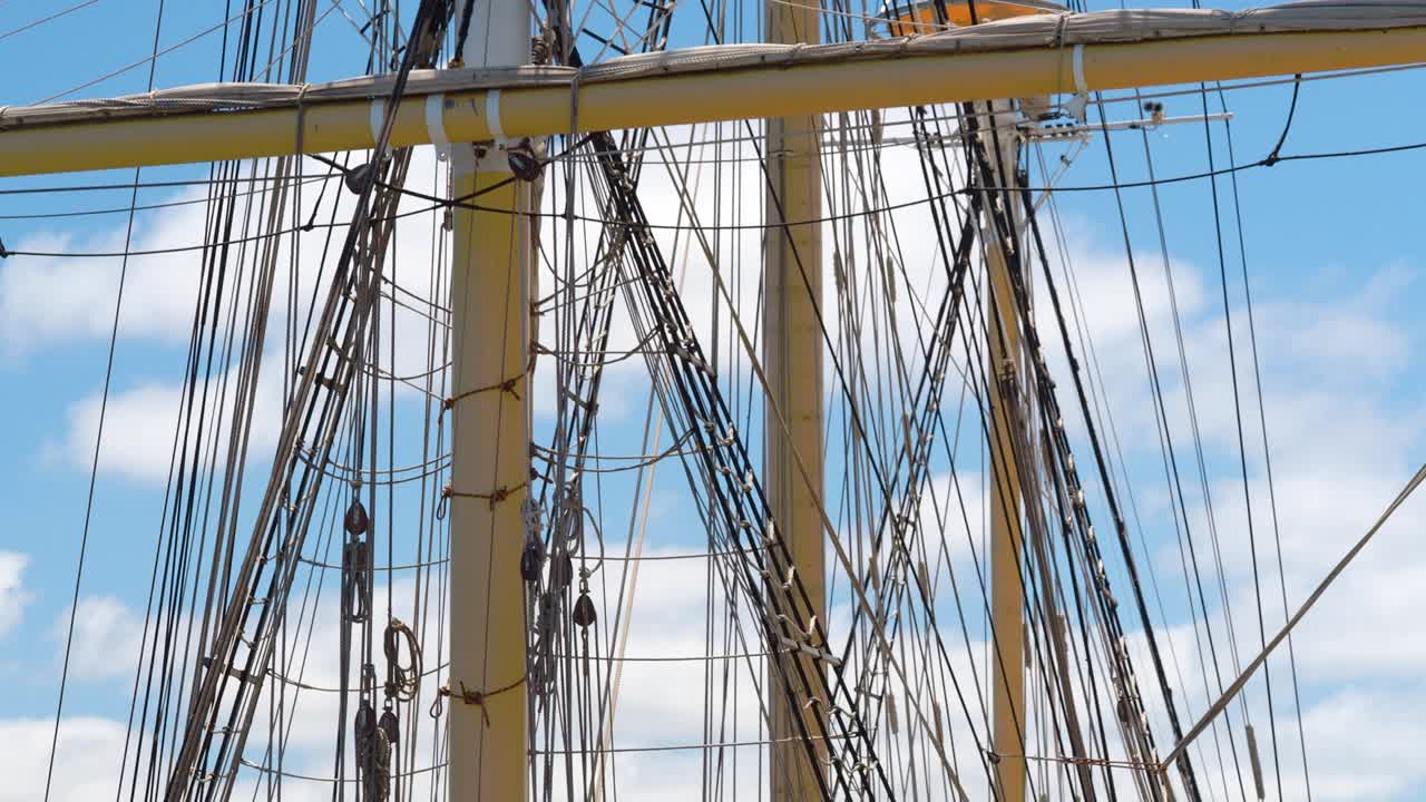 Tall ship rigging gently swaying, backlit against a blue sky with puffy clouds