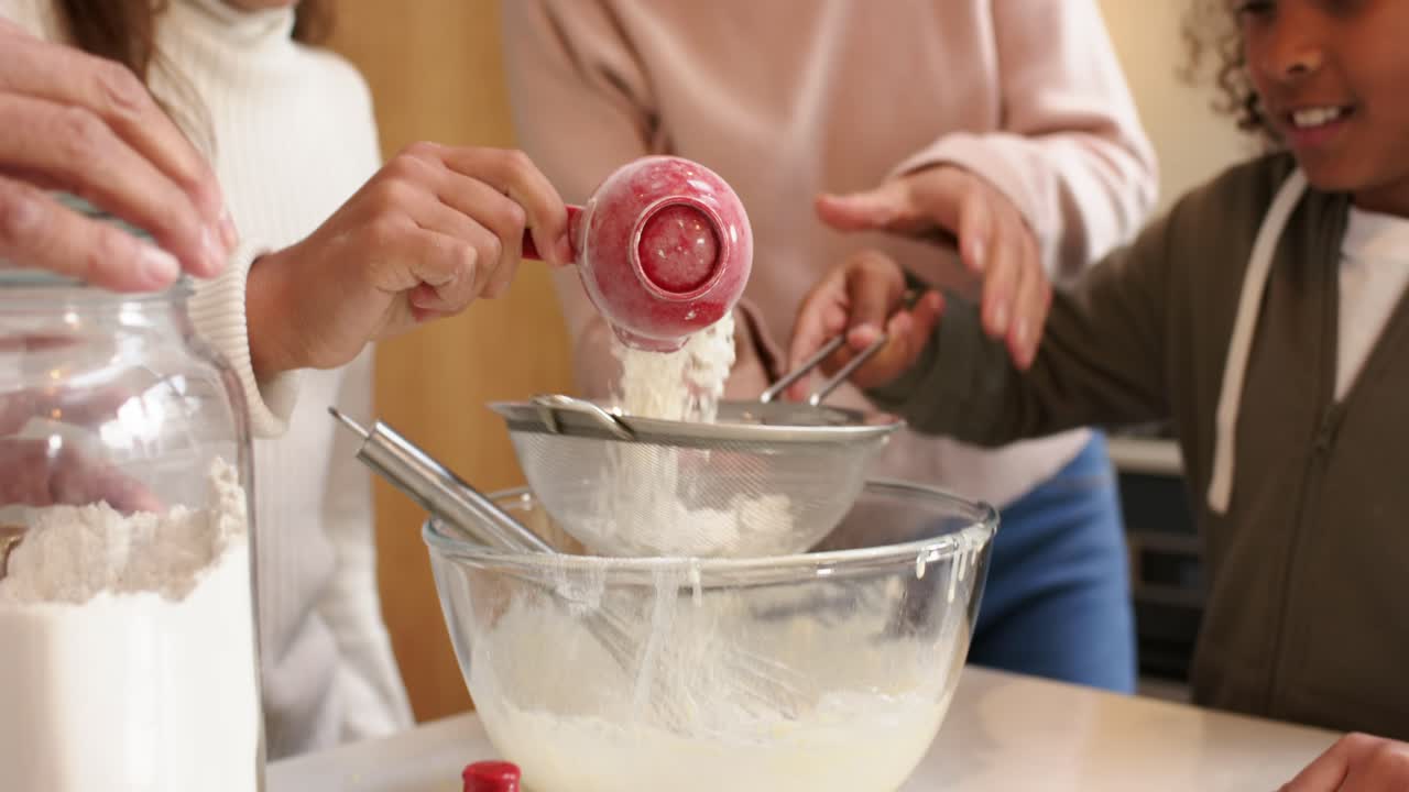 Keluarga memanggang, ibu yang memotivasi membimbing anak-anak mengayak tepung dengan saringan dan cangkir di dapur.