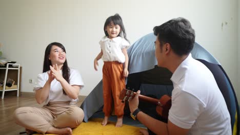 happy asian father playing ukulele guitar while little daughter and mother singing together in front of tent at home