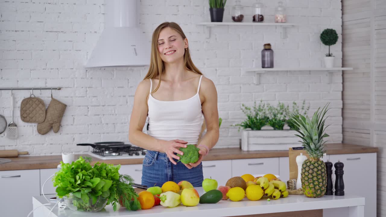 Cheerful female with broccoli at home. Sportive young woman choosing healthy diet full of minerals and vitamins in the kitchen. Girl standing near fresh fruit and vegetables.