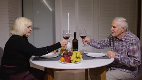 Portrait-of-senior-old-couple-toasting-wine-at-home-during-romantic-supper-in-kitchen-at-home