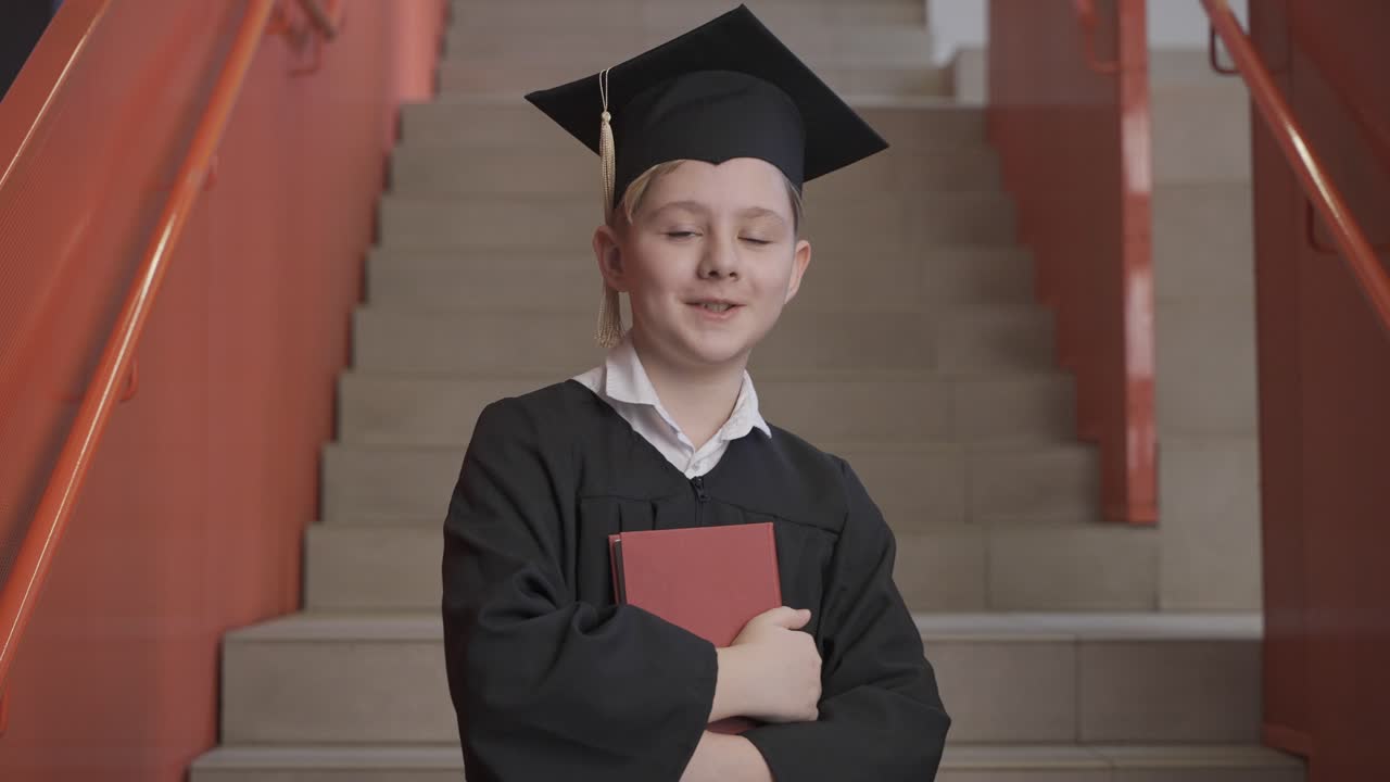 Portrait of a happy Caucasian preschool male student in cap and gown holding book and looking at the camera at the graduation ceremony