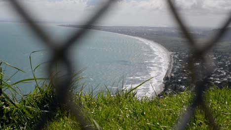 grassy edge of a mountain towering above a oceanfront town with chain-link fence in foreground