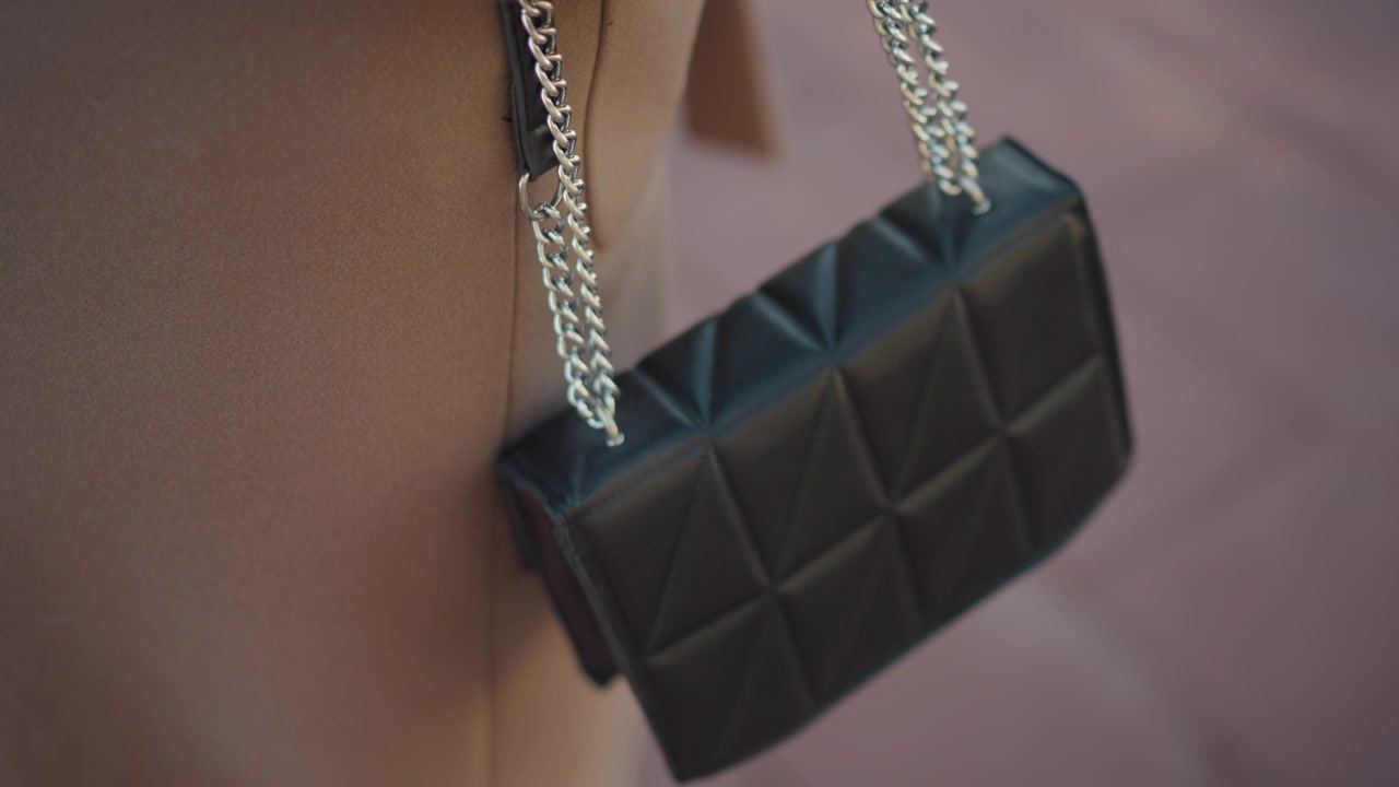 Primo piano di una borsa in pelle trapuntata nera con tracolle a catena argentate, appesa al braccio. La borsa presenta cuciture geometriche e un'illuminazione soffusa ne evidenzia i dettagli della texture.