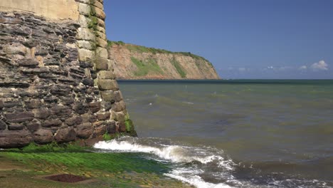 Small-waves-crashing-on-slippery-boat-ramp-on-north-yorkshire-coast-with-blue-skies,-sun-shining-and-large-steep-cliff-side-in-the-background