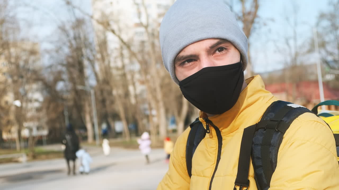 Food delivery man in a park looking into camera. Black medical mask, yellow backpack and jacket. Winter