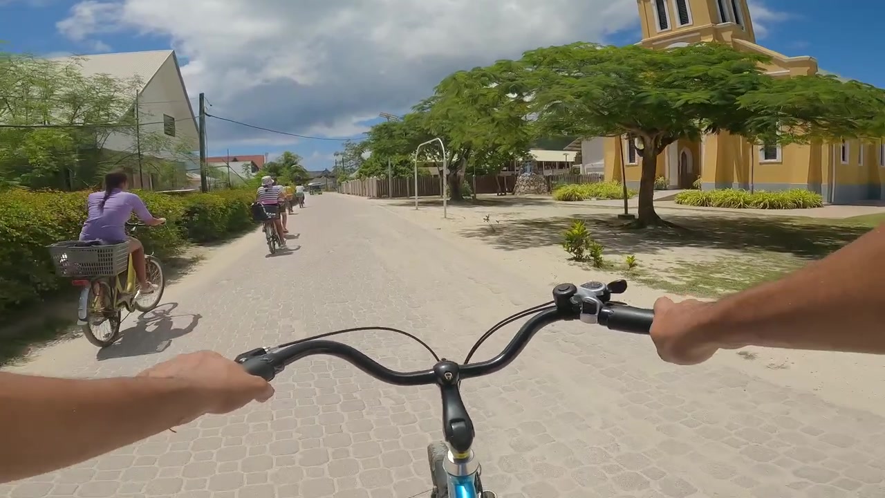POV andando de bicicleta pela icônica igreja católica amarela na Ilha La Digue, Seicheles. Com família de bicicleta, em um dia ensolarado, 50fps 013.