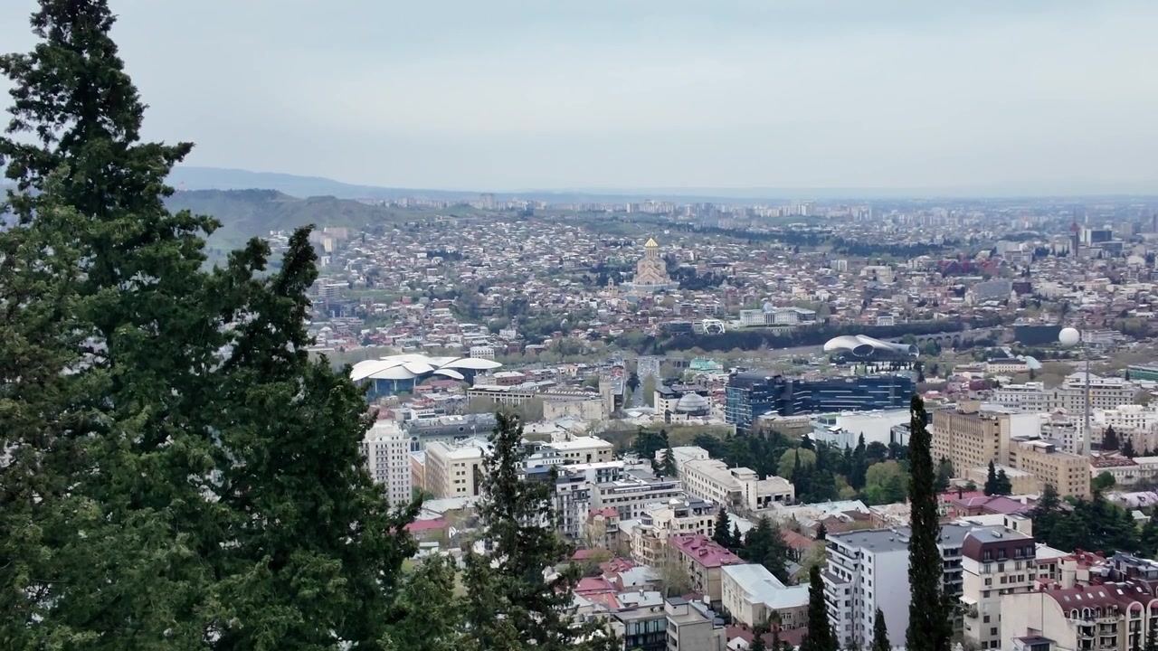Rozsáhlý širokoúhlý pohled na rozlehlou městskou krajinu Tbilisi pod zataženou oblohou. Stromy na levé straně rámují výhled, odhalující nespočet budov a vzdálených památek.