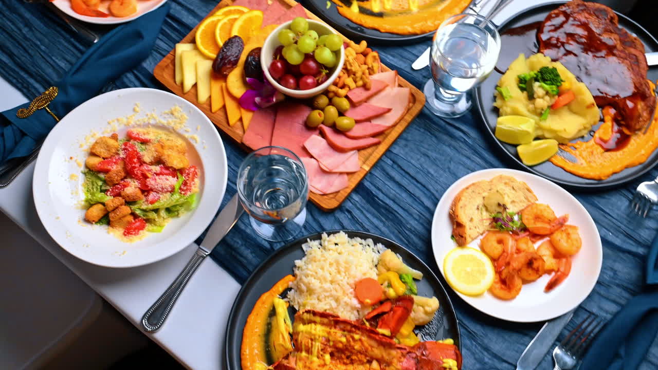 Vista dall'alto di un assortimento di cena mediterranea e a base di pesce. Composizione piatta di vari piatti tra cui bistecca, aragosta, insalata, gamberi e antipasti di formaggio su una tovaglia blu testurizzata.