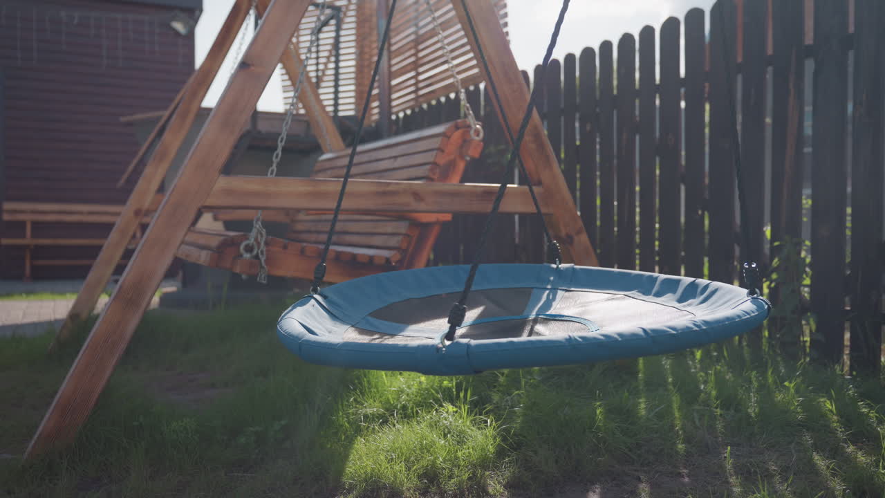 Close up of backyard playground with wooden bench swing and round blue nest swing gently suspended above grassy ground, sunlit by warm daylight, beside tall wooden fence and slatted shade panels