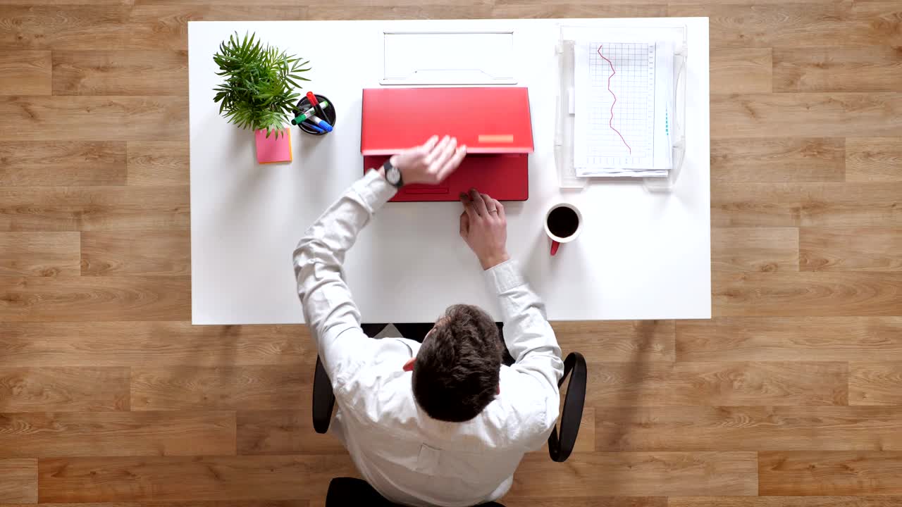 Young man walking in with coffee, sitting down and opening laptop, topshot, sitting behind desk