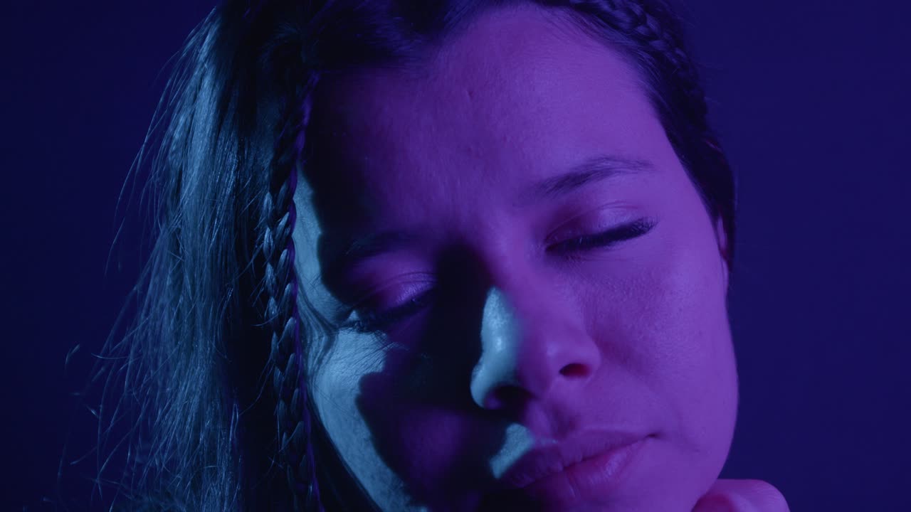 Engaging close-up of woman with braided hair, illuminated by vibrant, moody purple and blue lighting. Captivating, intimate atmosphere highlights expressive details