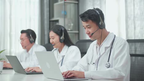 three asian doctors with stethoscopes wearing headsets working as call center agents sitting on the chairs and typing on computers while speaking to customers on the call at the office