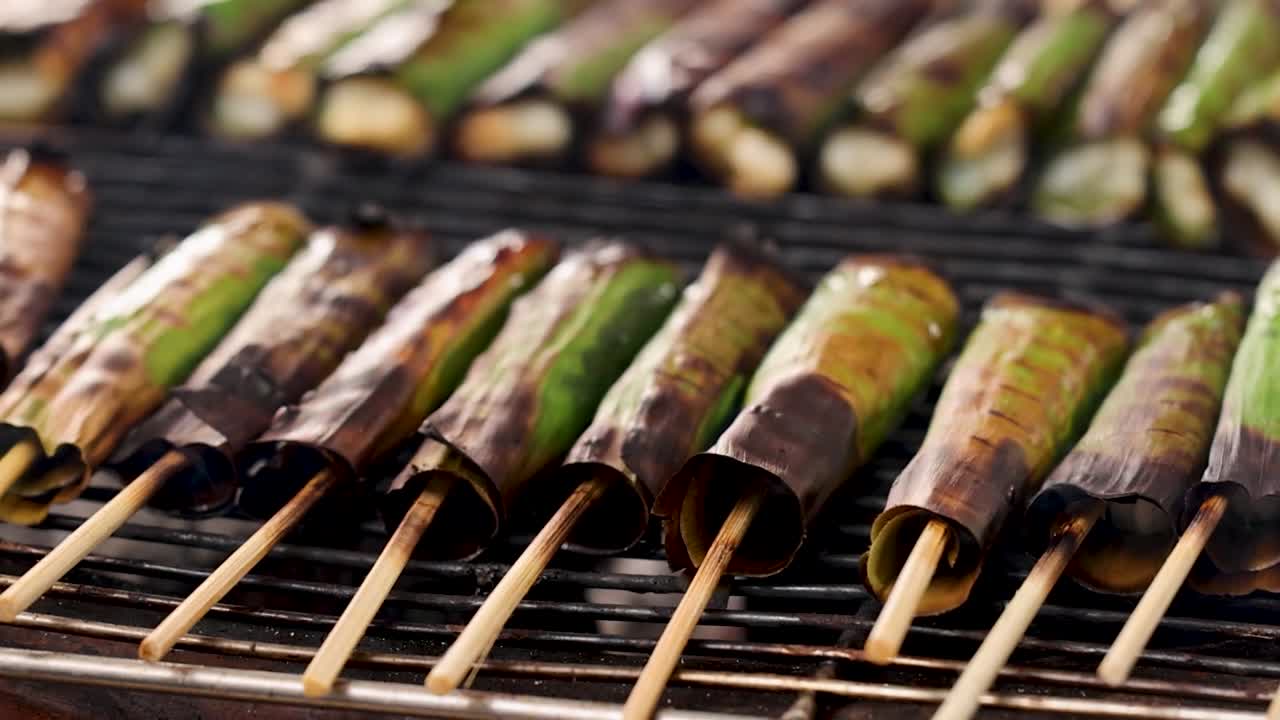 Primo piano di spiedini avvolti in foglie di banana che grigliano su un barbecue.