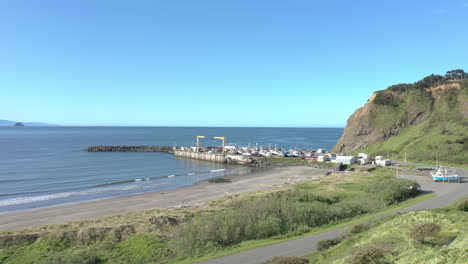 Drone-flying-low-over-grass-towards-boat-dock-in-Port-Orford,-Oregon-Coast,-during-a-summer-morning-with-blue-sky