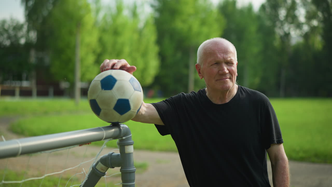 An elderly man in a black top rests his hand on a soccer ball placed on a goalpost, he stands calmly with, with a blurred view of a paved road and lush green grass